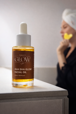 Daily Glow Up Gua Sha Glow Facial Oil im Fokus mit Gua Sha Anwendung im Hintergrund.